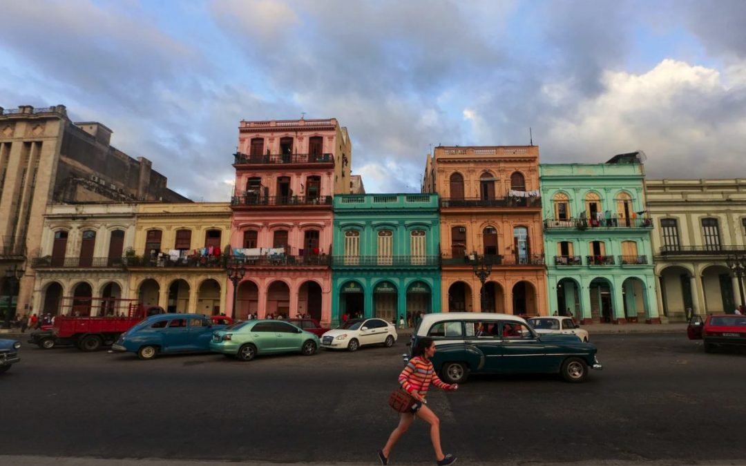 Calle de La Habana con edificios de fachadas de colores, autos clásicos estacionados y una mujer cruzando la calzada al atardecer